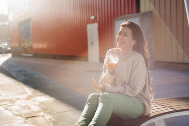 Fröhliche junge Frau, die ihr Getränk genießt, während sie auf einer Bank vor einem Einkaufszentrum sitzt - JMPF00321