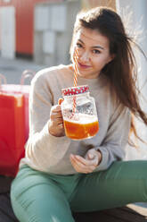 Lächelnde junge Frau zeigt Saft in einem Einmachglas, während sie im Sonnenlicht sitzt - JMPF00323