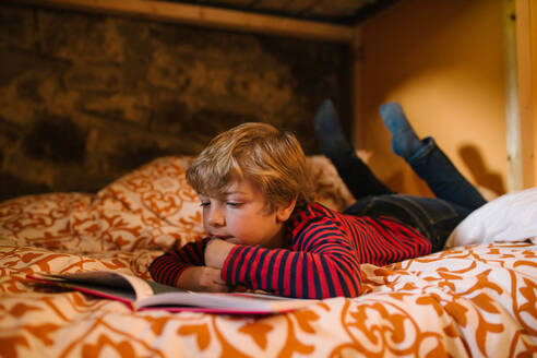 Dreamy child in casual wear lying on cozy bed and enjoying interesting fairy tale in story book for children - ADSF10708