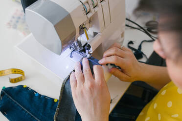 From above cropped unrecognizable brunette adult woman using sewing machine to make denim garment while working in home workshop - ADSF10713