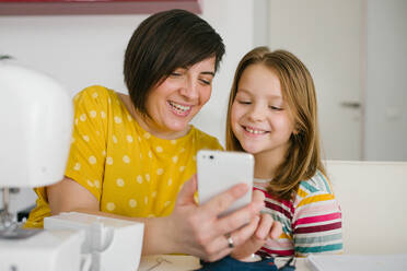 Cheerful adult woman smiling and taking selfie with girl while sitting at table and working in dressmaking workshop at home - ADSF10714