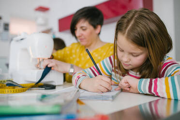 Konzentriertes Mädchen, das Hausaufgaben macht, während es neben einer erwachsenen Frau sitzt, die zu Hause ein Kleidungsstück näht - ADSF10715