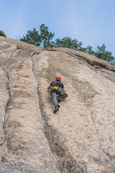Von unten anonymer Mann, der in der Natur mit Kletterausrüstung auf einen Felsen klettert - ADSF10722