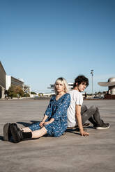 Attractive sensual blond woman looking at camera sitting with boyfriend on skateboard and dreaming against blue sky and blurred modern buildings on square - ADSF10741