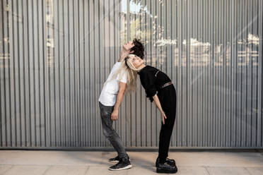 Seitenansicht eines trendigen Paares, das sich gegenseitig unterstützt, während es Rücken an Rücken steht und gegen die gestreifte Wand eines modernen Gebäudes träumt - ADSF10748