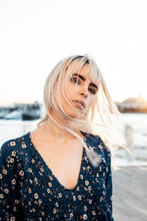 Gorgeous pensive blond female with black eyebrows in blue oversize dress looking at camera with curiosity while standing and touching hair on street during sunset - ADSF10754