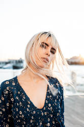 Wunderschöne nachdenkliche blonde Frau mit schwarzen Augenbrauen in blauem Oversize-Kleid, die neugierig in die Kamera schaut, während sie auf der Straße bei Sonnenuntergang steht und ihr Haar berührt - ADSF10754