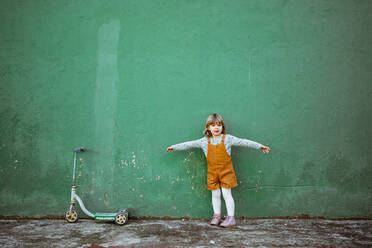 Little girl with outstretched arms standing near weathered green wall and kick scooter and counting while playing hide and seek - ADSF10770