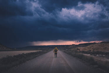 Rückenansicht eines jungen Mannes in gelber Jacke und Rucksack, der auf einer leeren Straße steht und sich hoch zu den düsteren, abendlichen, atemberaubenden Wolken in der Halbwüste Bardenas Reales Navarra Spanien trägt - ADSF10776