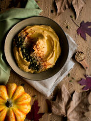 Set aus getrockneten Herbstblättern und Stoffservietten auf einem Holztisch neben einer Schüssel mit leckerem Kürbis-Hummus mit Kernen und Meersamen - ADSF10794