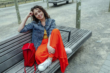 Smiling young woman sitting on bench and looking at camera - OGF00470