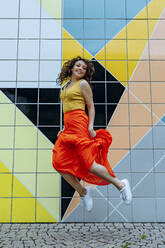 Young woman jumping in front of colorful tiled wall - OGF00480