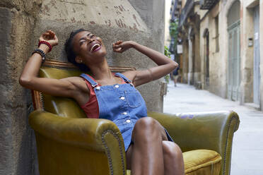 Fröhliche Frau mit erhobenen Armen auf einem alten Sessel auf der Straße sitzend - VEGF02647