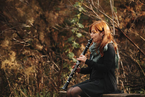 Schöne rothaarige Musikerin übt Klarinette spielen und sitzt auf einer Bank im Wald - MRRF00270