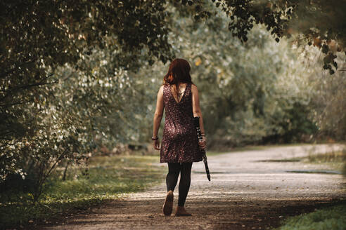 Young redhead woman walking with clarinet on footpath in forest - MRRF00275