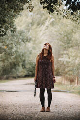 Smiling young woman holding clarinet while looking up at tree and standing on footpath in forest - MRRF00278