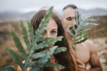 Woman and man hiding face with leaves in field during sunset - MIMFF00172