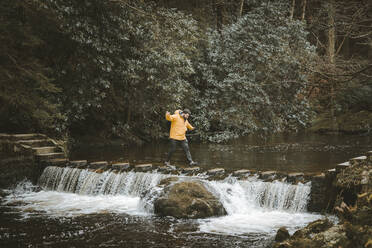 Seitenansicht eines männlichen Touristen in leuchtend orangefarbener Jacke, der auf einem Steg geht und einen Fluss überquert, durch den Wasser durch Trittsteine in einem Wald in Nordirland fließt - ADSF10813