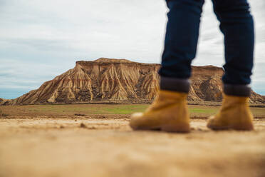 Beine eines gesichtslosen Reisenden in braunen Stiefeln und blauen Jeans, der auf einer schmutzigen Sandstraße mit Bergen und Himmel im unscharfen Hintergrund in Bardenas Reales, Navarra, Spanien steht - ADSF10848