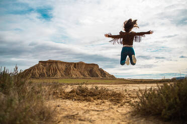 Rückansicht einer nicht erkennbaren Dame in stilvoller Freizeitkleidung, die ihre Arme im Sprung in einem verlassenen Tal mit großen braunen Klippen und blauem Himmel im Hintergrund in Bardenas Reales, Navarra, Spanien ausbreitet - ADSF10849