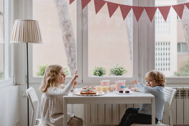 Seitenansicht von fröhlichen kleinen Kindern in Freizeitkleidung, die an einem Holztisch in der Nähe des Fensters sitzen und süßen Kuchen in den Ferien essen - ADSF10852