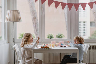 Side view of cheerful little kids in casual clothes sitting at wooden table near window and eating sweet cake during holiday - ADSF10852