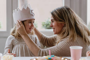 Seitenansicht einer fröhlichen Mutter, die ihrer Tochter eine handgefertigte Krone aus Filz aufsetzt, während sie gemeinsam zu Hause Geburtstag feiert - ADSF10853