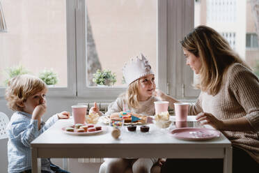 Positive mother in casual clothes sitting at wooden table and talking with kids celebrating birthday at home - ADSF10854