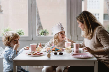 Positive Mutter in Freizeitkleidung sitzt am Holztisch und spricht mit Kindern, die zu Hause Geburtstag feiern - ADSF10854