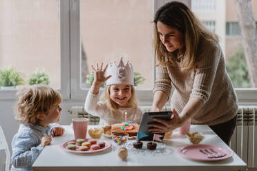 Positive Mutter und Tochter in Freizeitkleidung sitzen zusammen am Tisch und führen einen Videoanruf auf einem Tablet durch, während sie zu Hause Geburtstag feiern - ADSF10856
