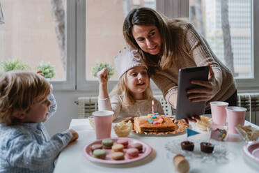 Positive Mutter und Tochter in Freizeitkleidung sitzen zusammen am Tisch und führen einen Videoanruf auf einem Tablet durch, während sie zu Hause Geburtstag feiern - ADSF10857