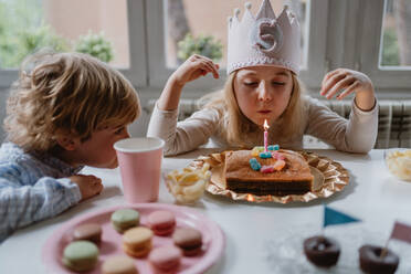 Glückliches kleines Mädchen in Freizeitkleidung und mit Filzkrone bläst eine Kerze auf dem Geburtstagskuchen aus, während sie an einem Holztisch sitzt - ADSF10860