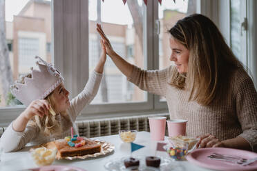 Nettes kleines Mädchen in Freizeitkleidung und Papier Krone geben High Five mit Mutter, während am Tisch sitzen und essen leckeres Essen während der Ferien - ADSF10861
