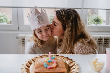 Liebevolle Mutter, die ihr kleines Mädchen küsst und beglückwünscht, während sie bei einer Geburtstagsfeier am Tisch zu Hause Zeit miteinander verbringt - ADSF10862