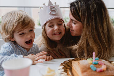 Liebevolle Mutter und Bruder, die einem kleinen Mädchen gratulieren, während sie bei einer Geburtstagsfeier am Tisch zu Hause Zeit miteinander verbringen - ADSF10864