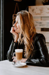 Seitenansicht einer langhaarigen, trendigen, schönen blonden Frau, die in einem Café sitzt und aus einem Glas köstlichen, schaumigen Kaffees trinkt - ADSF10876