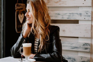 Seitenansicht einer langhaarigen, trendigen, schönen blonden Frau, die in einem Café sitzt und aus einem Glas köstlichen, schaumigen Kaffees trinkt - ADSF10877