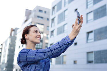 Cheerful female entrepreneur in stylish jacket smiling and using smartphone to take selfie - ADSF10897