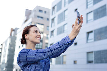 Fröhliche Unternehmerin in modischer Jacke, die lächelt und ein Smartphone für ein Selfie benutzt - ADSF10897