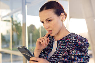 Cheerful female entrepreneur smiling and looking away while talking on smartphone and standing on background of modern buildings and cloudless blue sky - ADSF10900