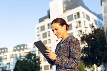 Serious businesswoman in elegant jacket browsing tablet and looking away while standing on city street on sunny day - ADSF10901