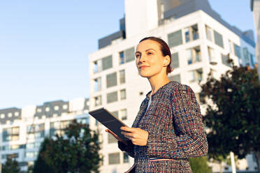 Serious businesswoman in elegant jacket browsing tablet and looking away while standing on city street on sunny day - ADSF10902