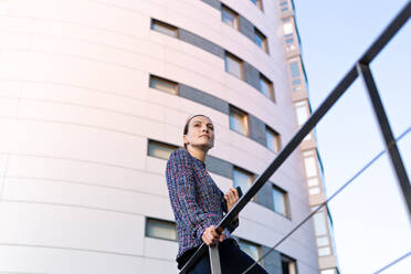 Low angle of serious female manager in elegant outfit leaning on railing and looking away while standing outside modern office building - ADSF10904