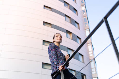 Niedriger Blickwinkel einer ernsthaften Managerin in elegantem Outfit, die sich an ein Geländer lehnt und wegschaut, während sie vor einem modernen Bürogebäude steht - ADSF10904