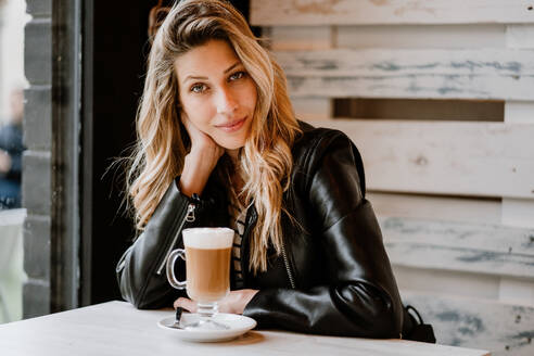 Seitenansicht einer langhaarigen, trendigen, schönen blonden Frau, die in einem Café sitzt und in die Kamera schaut, während sie aus einem Glas köstlichen, schaumigen Kaffees trinkt - ADSF10905