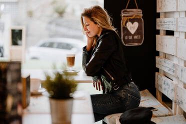 Side view of long haired trendy beautiful blonde woman sitting in a cafe shop drinking from a glass of delicious foamy coffee with closed eyes - ADSF10906