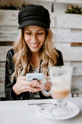 Stilvolle langhaarige Blondine in schwarzer Lederjacke und trendiger Mütze lächelt beim Surfen mit dem Handy in der Hand am Tisch im Café - ADSF10907