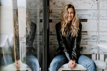 Trendy long haired woman in leather jacket and cap sitting in a wooden bench and looking at camera - ADSF10914