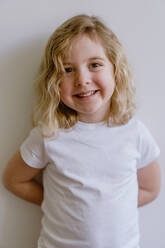 Positive child wearing casual clothes smiling and looking at camera while standing and leaning on white wall in modern studio - ADSF10919