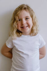 Positives Kind in Freizeitkleidung, das lächelt und in die Kamera schaut, während es steht und sich an eine weiße Wand in einem modernen Studio lehnt - ADSF10919